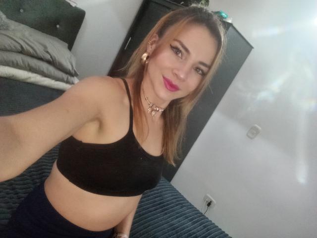 EmelySofia, con un seno di media grandezza, indossa una crop top nera, sorride alla fotocamera, ha il rossetto rosa, i capelli castano chiaro e una collana, ed è in piedi in una camera da letto..