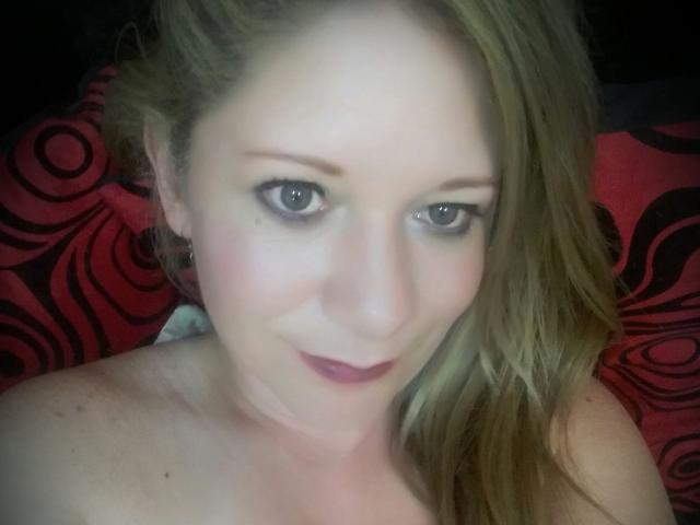 SecretTemptationX with pale skin, blue eyes, and blonde hair, looking up with a slight smile, wearing dark pink lipstick, and no visible clothes. Boobs partially visible.