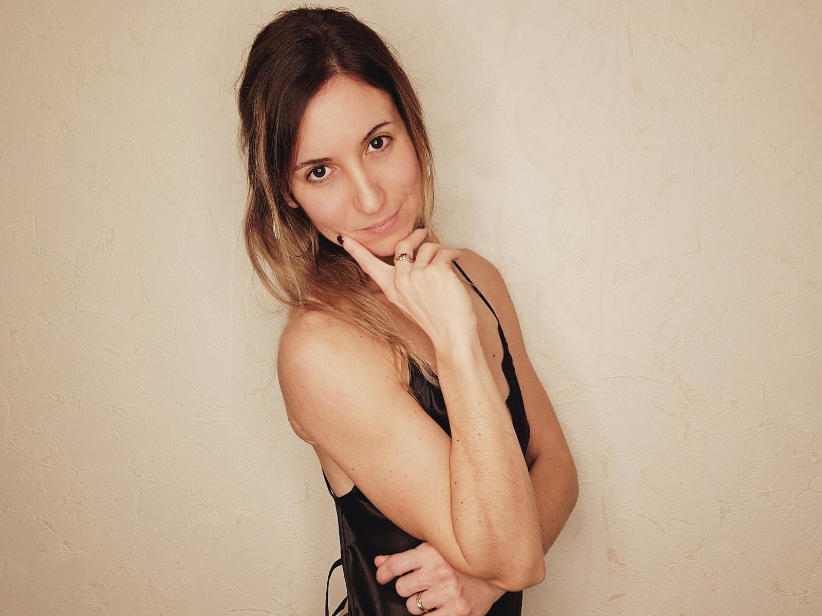 LexaDelMar with shoulder-length brown hair, wearing a black dress, touching her chin with a subtle smile, looking at the camera, her hands crossed over her chest.