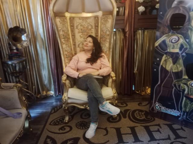 SakotoDika with wavy brown hair, pink sweatshirt, jeans, and white sneakers, sitting on an ornate gold and white throne, looking up with a relaxed expression.