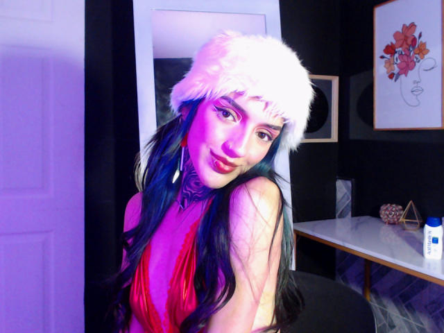 Amellie Jones, con cabello azul oscuro y largo, maquillaje rosa y un vestido rojo, lleva un sombrero blanco de piel y sonríe a la cámara. Hay luces moradas y azules, y se le ve el pecho. Está en una habitación..