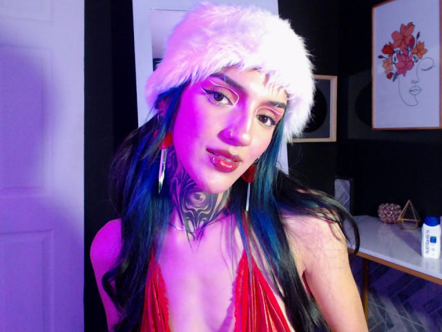 Amellie Jones met blauw-groen haar, een witte, pluizige kerstmuts, rode lippenstift, een getatoeëerde nek, een rode top met halter, paars-blauwe verlichting, grote oorbellen en een lichte glimlach. Borsten zichtbaar..