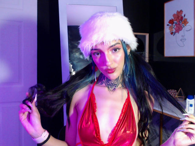 Amellie Jones, com longos cabelos azul-escuros, roupa íntima vermelha e brilhante, gorro de Papai Noel branco e peludo, segurando o cabelo, tatuagens, pulseira rosa, iluminação roxa, lábios rosados, paredes pretas, obras de arte emolduradas ao fundo..