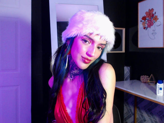 AmellieJones with long black hair, purple highlights, wearing a white fur hat, red velvet dress, and choker, smiling, showing her tits, in a purple-lit room.