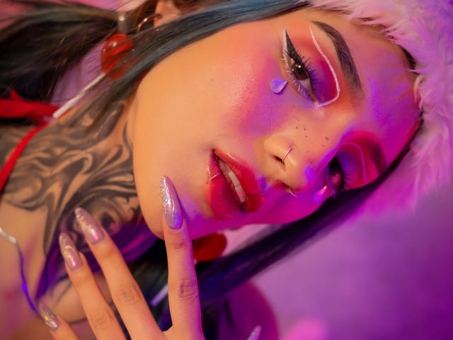 Amellie Jones, com cabelo rosa e azul, lábios vermelhos brilhantes, unhas com glitter, pescoço tatuado, piercing no nariz e maquiagem nos olhos, usando um chapéu branco e fofo, sob iluminação rosa e roxa..