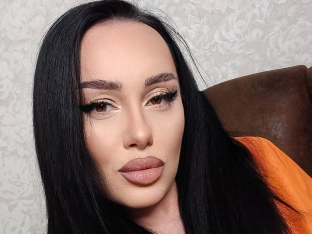 AnnaElyse, con cabello negro largo, ojos marrones y labios rosados y gruesos, lleva un delineador de ojos negro intenso y una blusa naranja, mirando ligeramente hacia un lado con una expresión neutra..