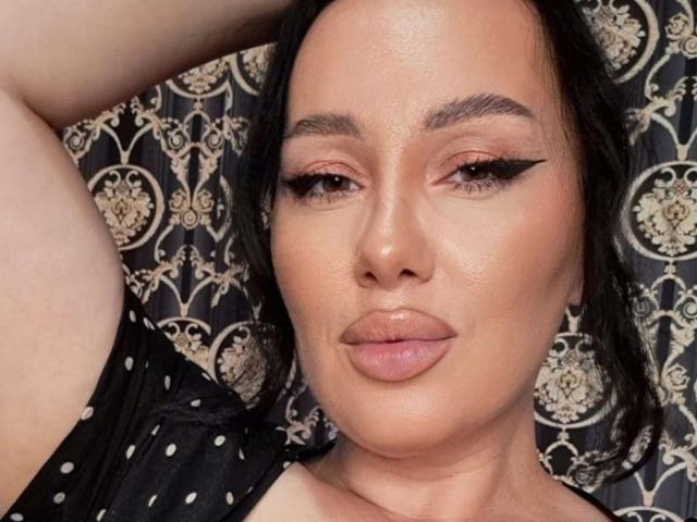 AnnaElyse with pouty, glossy pink lips, thick black eyeliner, and brown eyes. She's wearing a black, polka-dotted top, with her arm raised. Her skin is smooth and tan.
