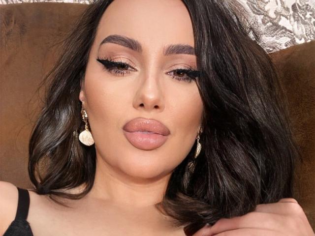 AnnaElyse tiene el cabello oscuro y ondulado, cejas pobladas, largas pestañas negras y labios gruesos de color rosado-marrón. Lleva aretes dorados y una blusa negra, con un fondo marrón..