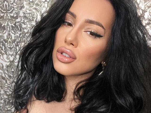 AnnaElyse has glossy black hair, brown eyes with winged eyeliner, and plump pink lips. Her skin is tan, and she wears small hoop earrings.
