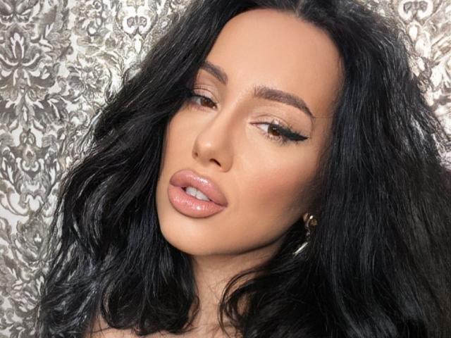 AnnaElyse tiene labios rosados y brillantes, cabello ondulado oscuro y ojos marrones con largas pestañas negras. Está mirando ligeramente hacia un lado con una expresión suave y sensual..