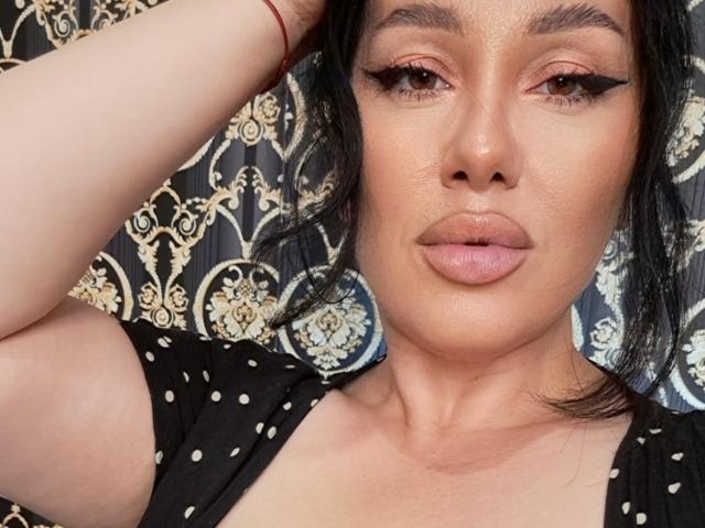 El rostro de AnnaElyse, mostrando su piel bronceada, delineador de ojos negro y labios carnosos, con un brazo levantado, vistiendo una blusa negra con lunares y una pulsera roja en la muñeca..