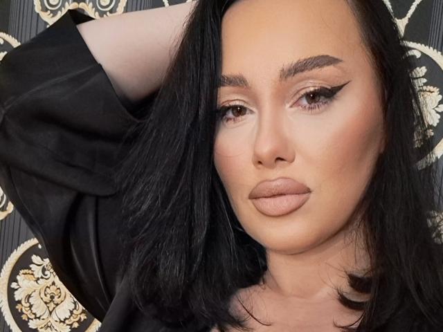 AnnaElyse, con cabello negro, brazo levantado, vistiendo una camisa negra, tiene un delineado de ojos alado pronunciado, ojos marrones y labios carnosos de color rosa-marrón, mirando directamente a la cámara..