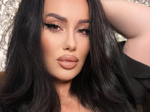 AnnaElyse with long black hair, full lips, and brown eyes, looking directly at the camera with a slightly raised arm. She's wearing black eyeliner and glossy pink lips.