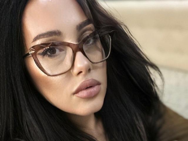 AnnaElyse with brown glasses, dark wavy hair, light skin, full pink lips, slightly parted, serious expression, looking slightly to the right.