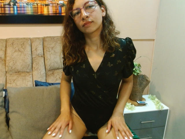VioletSingh with curly brown hair, glasses, black V-neck bodysuit, and white nails, sitting on bed, hands on thighs, showing her pussy through high-cut bodysuit.