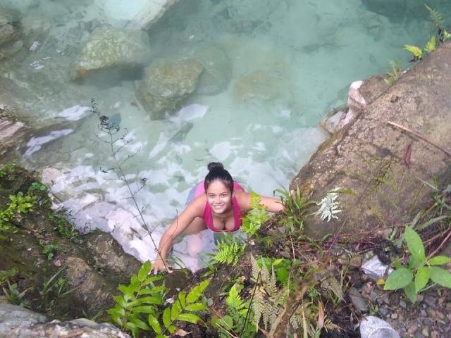 CrespaHot69 with big boobs, pink top, black hair in bun, smiling, kneeling in clear water, rocky shore, green plants, and ferns, holding rock, natural light, outdoor setting.