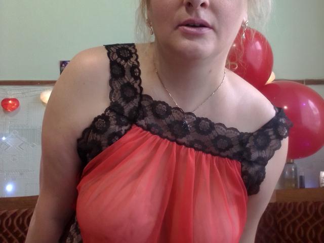 KelliRedhaer with her tits visible through a sheer red lace top, black lace trim, and a gold necklace, slightly open mouth, red balloons in the background.