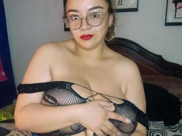 NaugthyCurvy, con lápiz labial rojo, gafas y una camiseta de malla negra, cubriéndose los pechos con una mano, tiene un cuerpo curvilíneo y de piel clara, y una expresión segura y sensual..