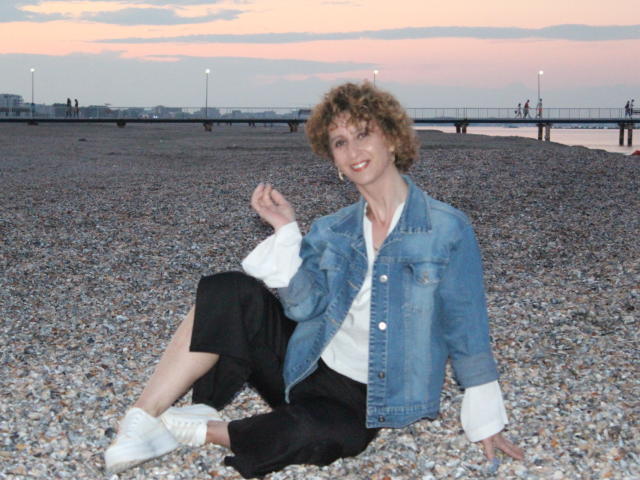 NikHotty with curly brown hair, blue denim jacket, white shirt, black pants, and white sneakers, sitting on pebbly beach, smiling, hand raised, sunset in background.