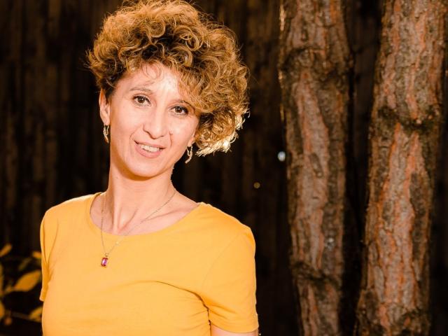 NikHotty with curly blonde hair, yellow top, small tits, smiling, wearing a pink pendant, standing in front of a tree, nighttime.