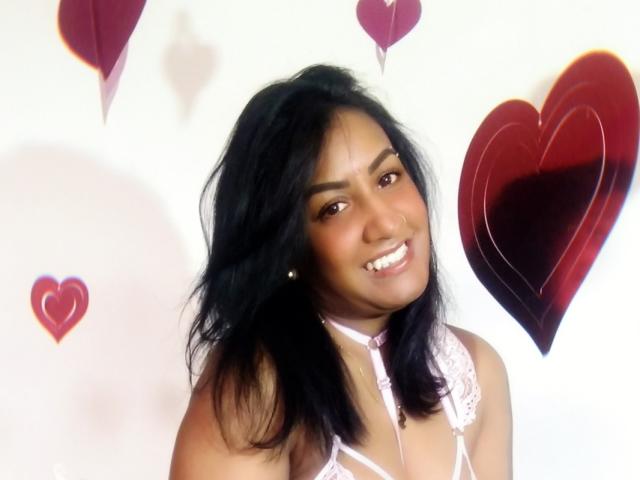 Nahirr has dark brown skin, black wavy hair, and is smiling with white teeth. She's wearing a pink lace bra and has small earrings. Her tits are visible.