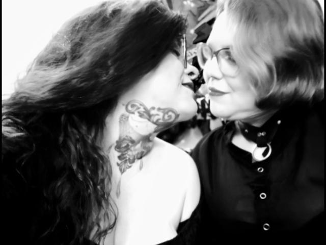 Fotografia in bianco e nero di due ragazze latine che si baciano, che mostra il loro seno prosperoso e i complessi tatuaggi sul collo, mentre una donna più anziana con gli occhiali e una camicia scura le osserva..