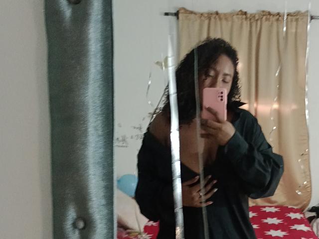 CaroldelaTorre mit lockigen Haaren, einem schwarzen, schulterfreien Oberteil und einem rosa Handy, macht ein Selfie vor einem Spiegel und zeigt dabei ihre kleinen Brüste. Im Hintergrund ist ein Bett mit einer roten Bettdecke mit Sternenmuster zu sehen..