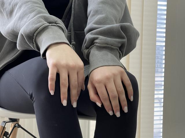 Le mani di MiaMoone, con le unghie curate, riposano sui suoi leggings neri, indossa una felpa grigia, è seduta su una sedia, e la luce proviene dalle persiane a destra..