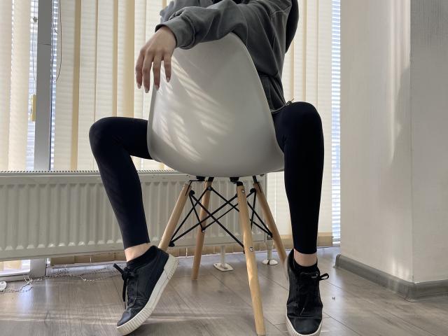 MiaMoone è seduta su una sedia bianca con gambe di legno, indossa una felpa grigia, leggings neri e scarpe da ginnastica nere, con i glutei leggermente sollevati e le mani appoggiate sulla sedia..
