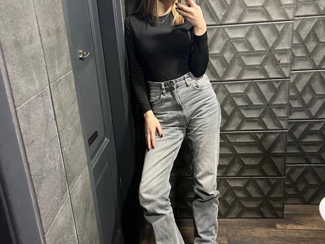 MiaMoone with a black long-sleeve top and high-waisted gray jeans, standing in a room, holding a phone, with a geometric patterned wall behind her.