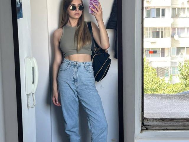 MiaMoone with long brown hair, wearing a green crop top, high-waisted light blue jeans, and aviator sunglasses, holding a phone with a pink case, standing by a wall.
