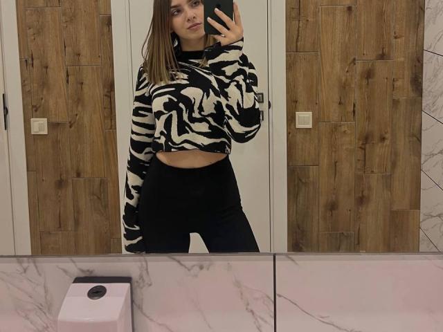 MiaMoone indossa un top corto a righe zebrate, che mostra il seno, e pantaloni neri a vita alta, e sta scattando un selfie allo specchio con il telefono, in piedi in un bagno con un mobile in marmo..