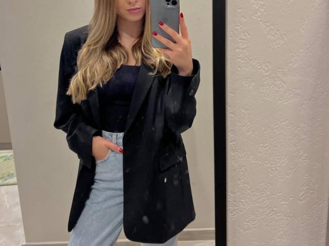 MiaMoone with long blonde hair, black blazer, black top, light grey high-waisted pants, red nail polish, holding phone, mirror selfie, hands in pockets, subtle smile.