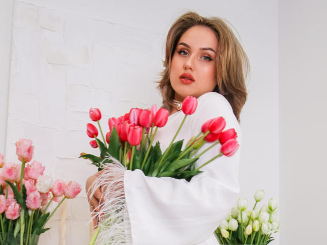 LisaFrancis with pink tulips in her hands, wearing a white shawl with fringes, has wavy brown hair and red lips, standing in front of white flowers and green stems.