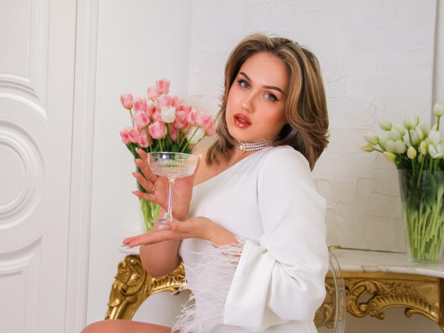 LisaFrancis with blonde hair, white dress, and choker, holding a glass, pink and white tulips, and white lilies in the background, sitting on an ornate gold chair.