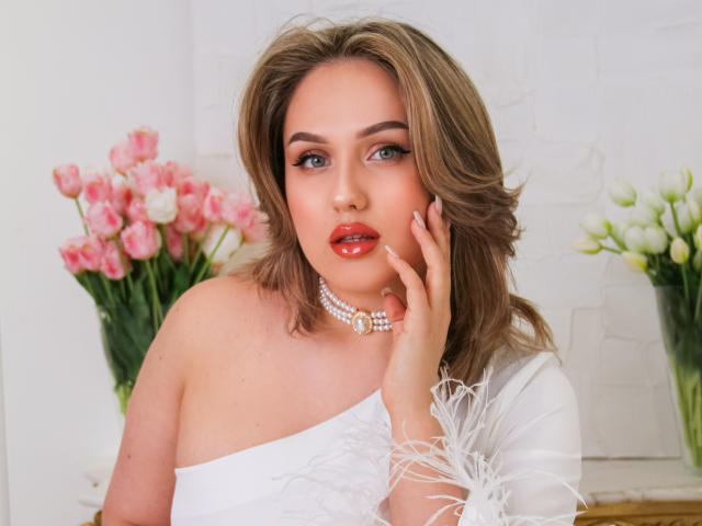 LisaFrancis with red lips, blue eyes, and brown wavy hair, wearing a white off-shoulder dress and pearl necklace, touching her cheek, surrounded by pink and white tulips.