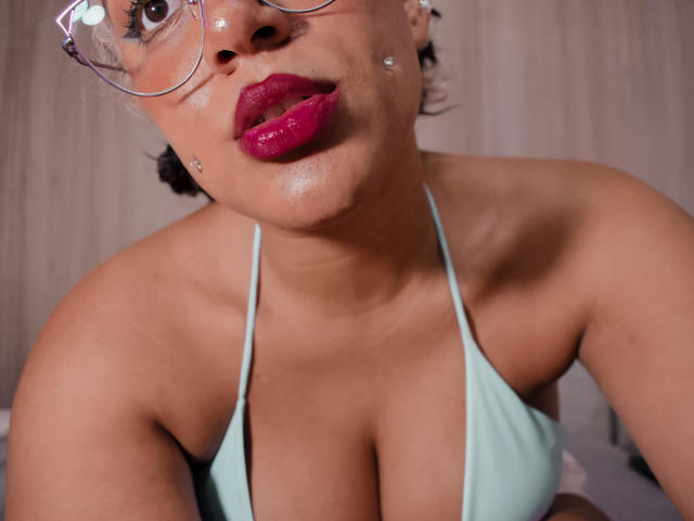 Natsukashii with shiny skin, red lipstick, glasses, light blue bikini top, showing her big tits, small silver stud on cheek, in a low angle shot.