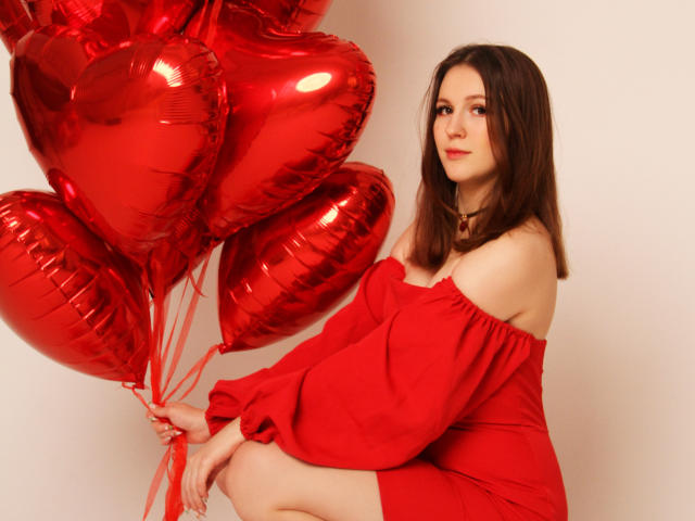 JuliaKing con globos rojos en forma de corazón, vestida con un vestido rojo de corte asimétrico, sentada, sosteniendo las cintas, mostrando sus pechos, cabello castaño, piel clara, labios rojos, fondo blanco..