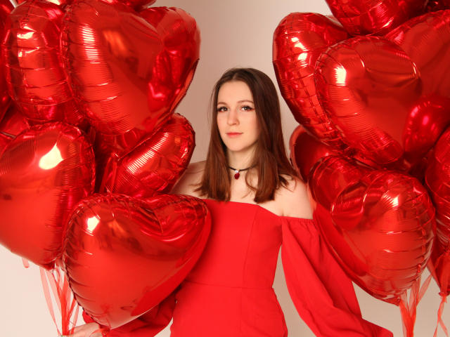 JuliaKing with red heart balloons, wearing a red off-shoulder dress, black choker, and red pendant, holding balloons, smiling, brown hair, white background, red balloons around.