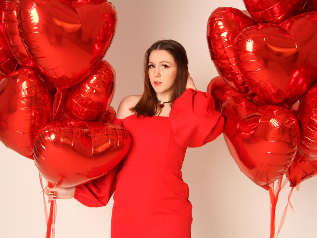 JuliaKing con globos en forma de corazón rojo, vestida con un vestido rojo de un solo hombro, sosteniendo dos globos a cada lado, con el cabello castaño suelto, fondo blanco, expresión seria..