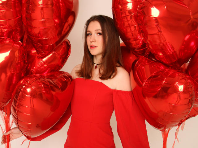 JuliaKing con un vestido rojo de un solo hombro, cabello castaño, globos rojos, usando un collar negro, mirando ligeramente hacia un lado, fondo blanco. Los senos parcialmente visibles, linda, sexy..