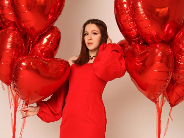 JuliaKing con globos rojos en forma de corazón, vestida con un vestido rojo de un solo hombro, sosteniendo globos a ambos lados, con una mano en el pelo, una cinta roja atada a los globos..