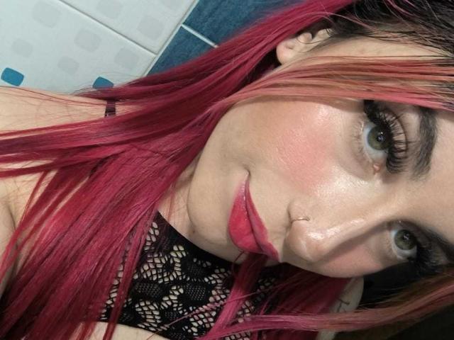 SammyViolet has pink hair, green eyes, and red lipstick, wearing a black lace top, with a nose ring, looking at the camera with a slight smile.