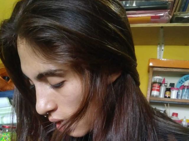 CalibanGray's profile, showing dark brown, shiny hair, nose piercing, and closed eyes, with a yellow wall and shelf in the blurred background. Her face is slightly tilted downward.