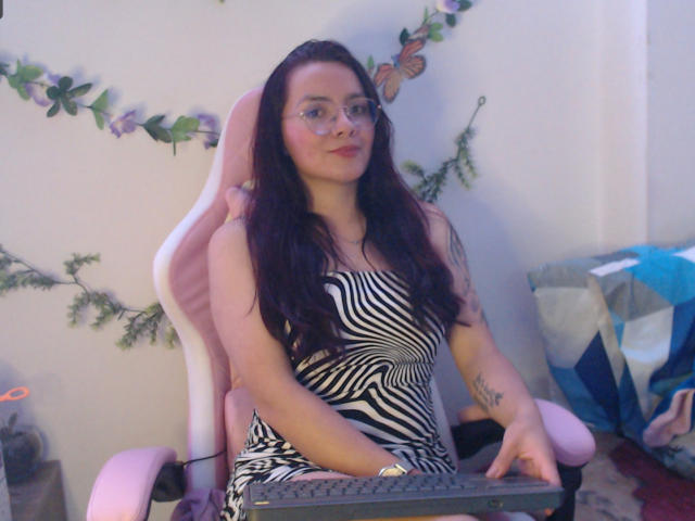 EimmyHoskX with long dark hair, glasses, zebra-striped dress, pink chair, keyboard, tattoo on right arm, butterflies on wall, smiling, holding keyboard, room with blue and gray blanket.