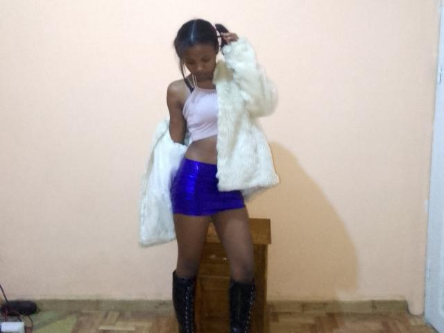 LunaHottiest with dark skin, wearing a white fur coat, pink tank top, shiny blue skirt, and black boots, standing in front of a wooden box.