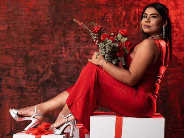 SusanClaire con vestido rojo, sosteniendo rosas, sentada en una caja, tacones plateados, cabello oscuro, piel bronceada, aretes, fondo rojo, mirando a la cámara, sexy, curvilínea, trasero, pechos, ramo en la mano..