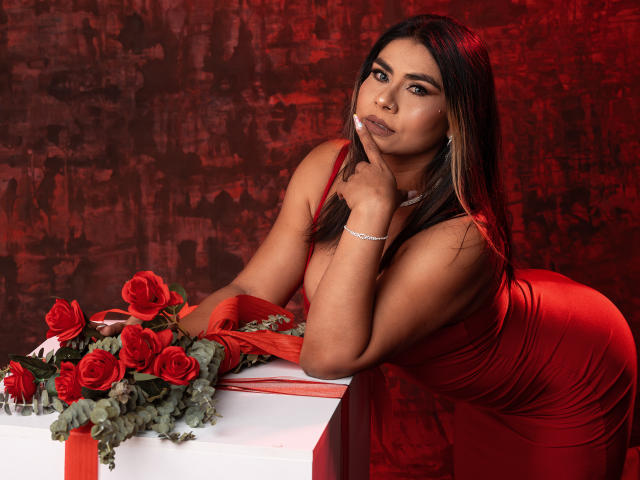 SusanClaire, vêtue d'une robe rouge, avec des cheveux bruns, et des roses rouges, appuyée sur une table blanche, un doigt sur le menton, regardant la caméra, un bracelet au poignet, fond texturé rouge et noir..