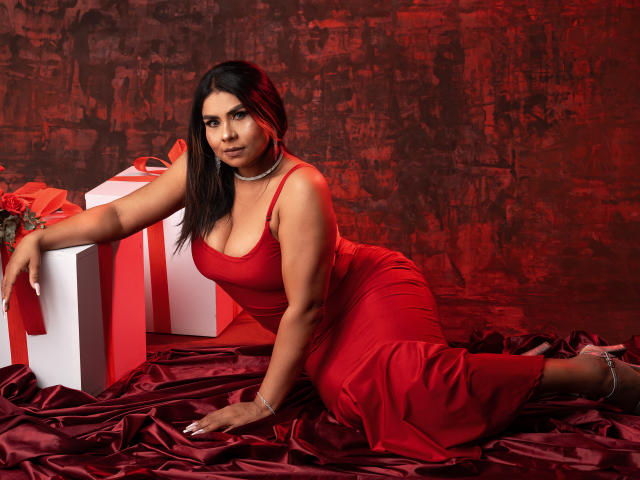SusanClaire mit großen Brüsten in einem roten Kleid, liegt auf rotem Satin, ein Arm auf einer weißen Geschenkbox, rotes Band, roter Hintergrund, schwarzes Haar, blickt in die Kamera, Perlenkette..