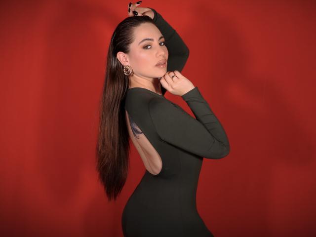 LuciaPerth with long brown hair, wearing a green long-sleeve dress, backless, showing a tattoo on her side, arms raised, looking over her shoulder, red background.